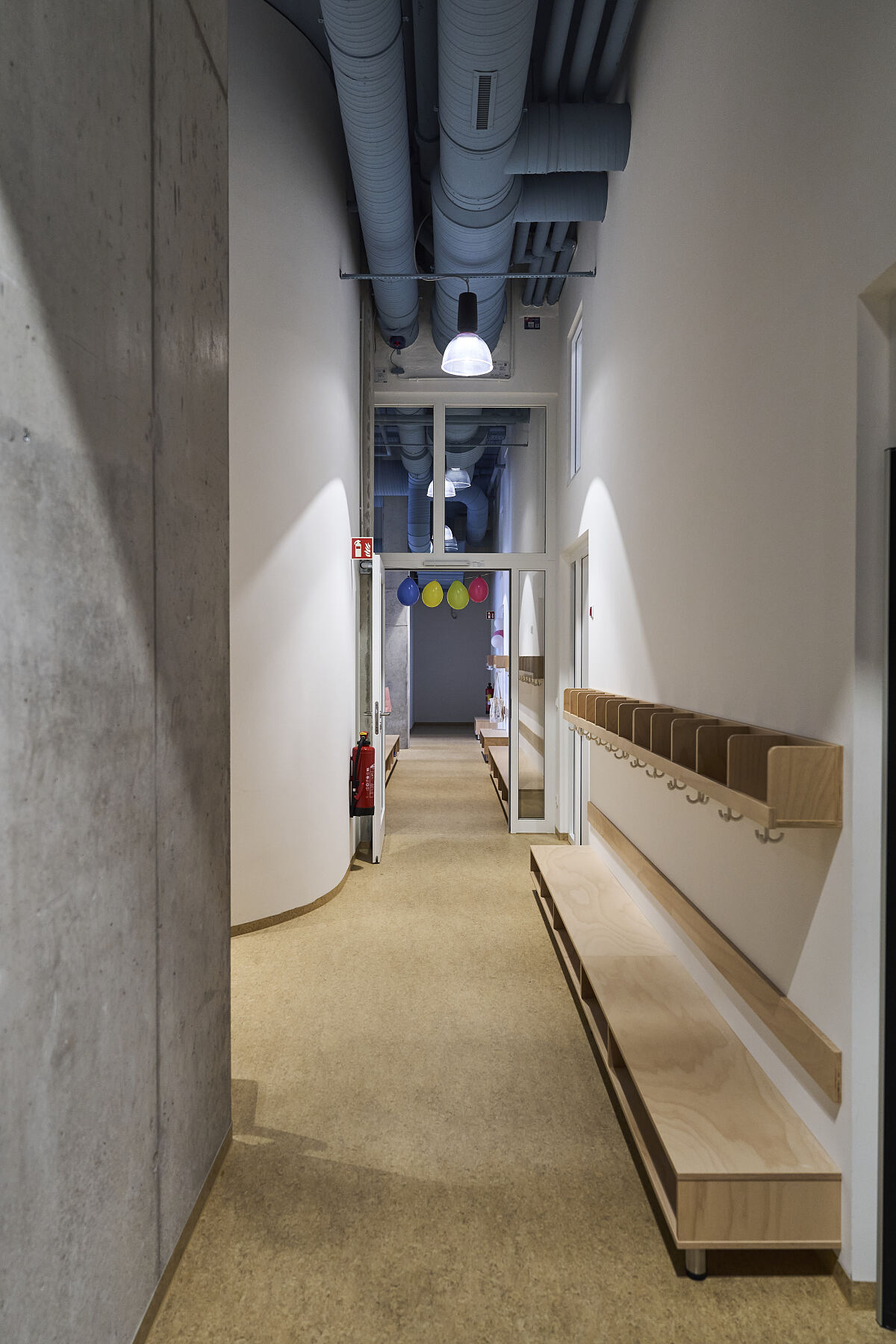 Blick durch den zentralen Garderobengang der neuen Kita. Kindgerechte Sitzbänke und Ablagen treffen auf die sichtbaren Leitungen und Betonoberflächen des Gebäudes M5.